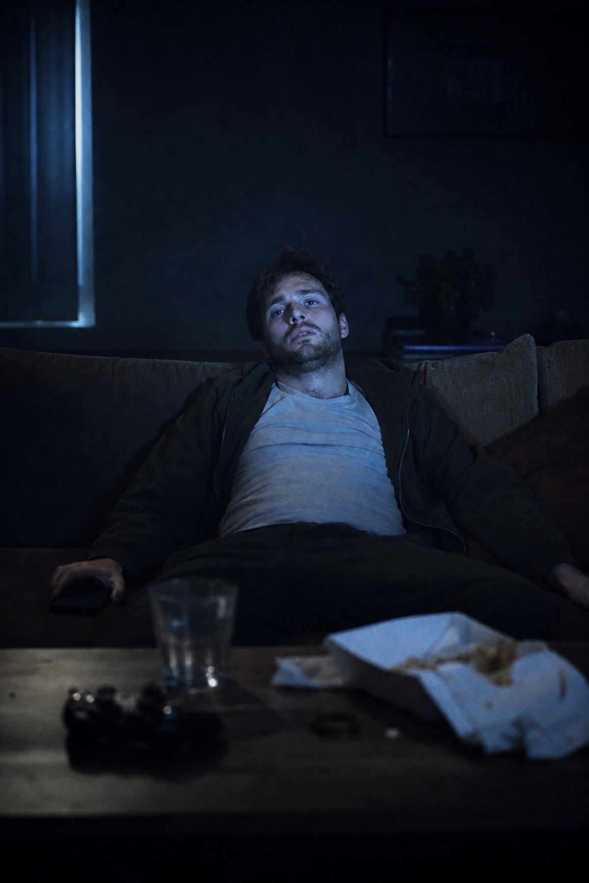A man slumped on a couch late at night, lit only by a television, staring at nothing.