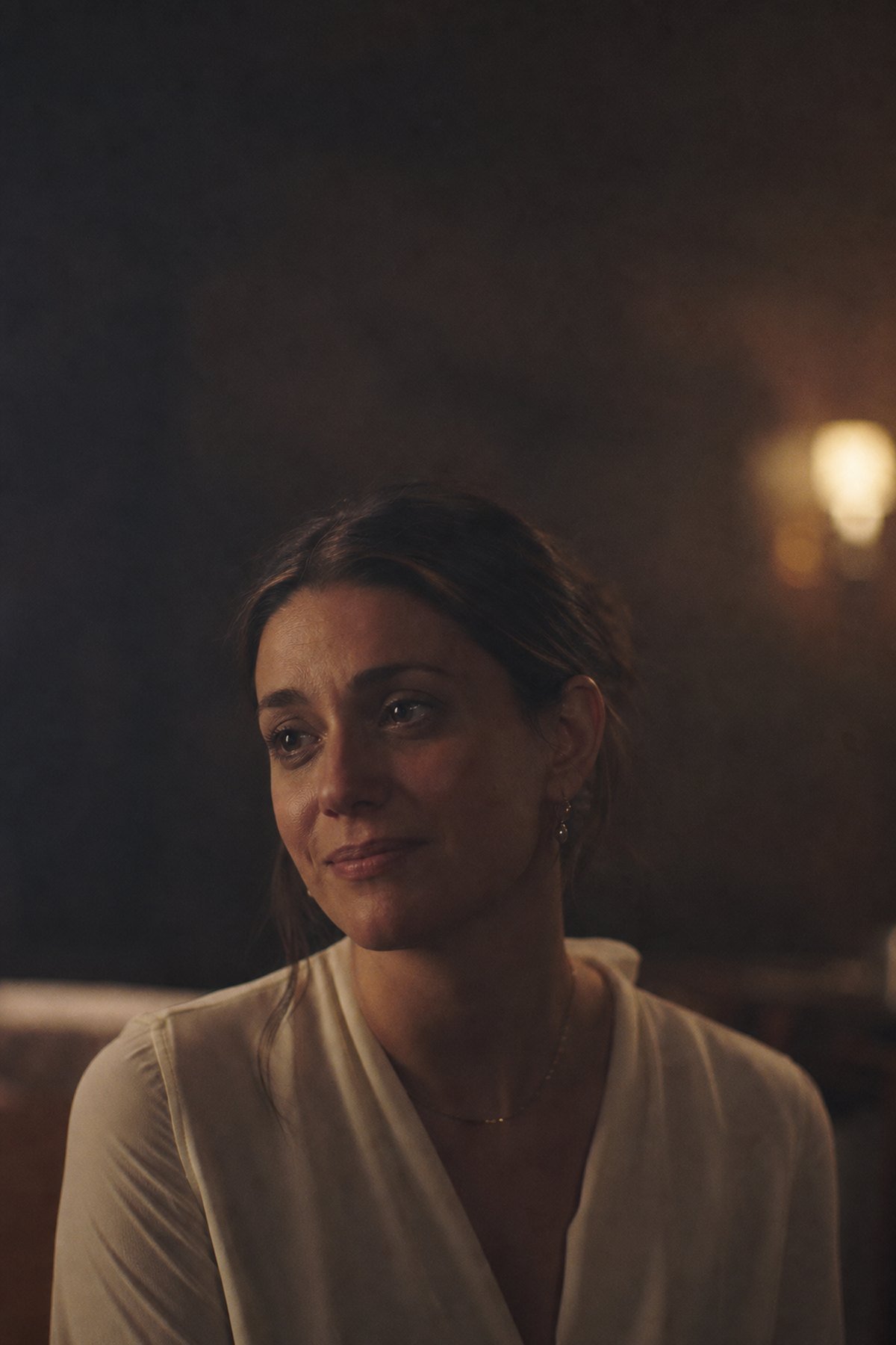 A woman sitting quietly in warm lamplight, tired and composed.