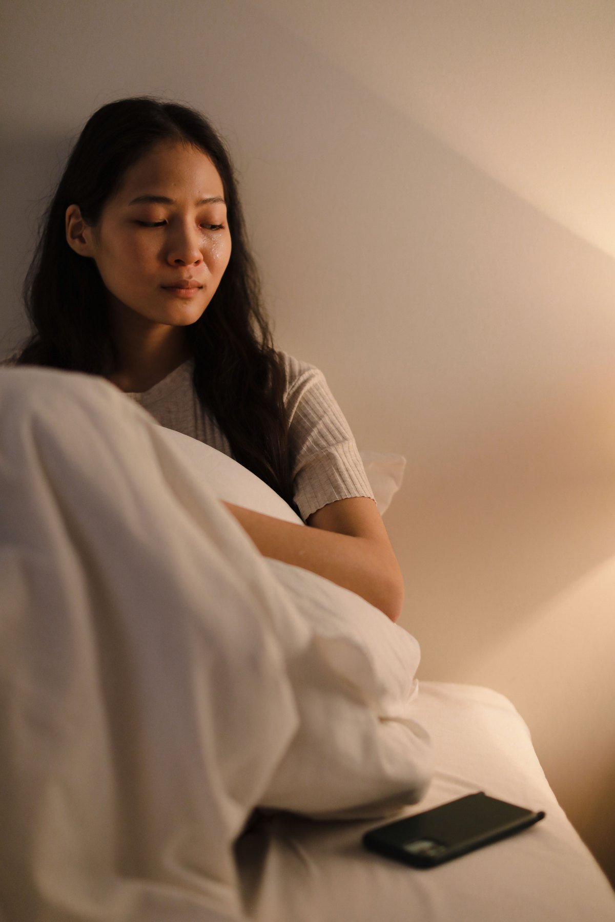 A woman sitting up in bed in warm lamplight, holding the duvet, looking down and away from the phone lying beside her.